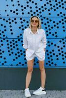 Slender young blonde woman in a white suit makes grimaces and has fun in summer against a blue wall photo