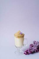 Beautiful dalgona drink a frothy coffee in a transparent mug and flowers lilac to purple Il purple background. Next to the mug is a marshmallow and a branch of lilac. photo