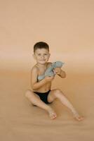 A child boy in underpants holds a soft bear toy in his hands, sitting on a beige background in the studio and smiling photo