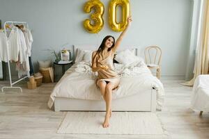Cute woman with long hair is sitting on a bed in a cozy bedroom at home with a cake on a plate and a candle and celebrating her birthday photo