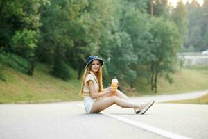 A happy girl in white shorts and a yellow blouse holds a glass of juice with a straw in her hands and sits on the road near the forest photo