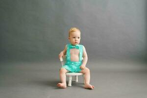 A funny little boy in a summer suit is sitting on a white children's chair on a gray background in a photo studio