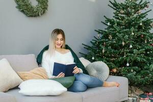 hermosa mujer es leyendo un libro, descansando en almohadas con un cobija a el Navidad árbol a hogar. foto