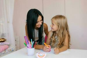 Smiling mother helps her adorable daughter with homework at room photo