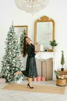 retrato de un hermosa joven sonriente mujer en un negro vestir con lentejuelas, participación un regalo en su manos. celebrando Navidad en el vivo habitación por el Navidad árbol y hogar foto