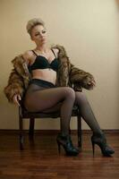 Attractive blonde model with pantyhose, black bra and fur coat posing provocatively on chair. Fashion portrait of sensual blonde, studio shot. Sensual female in black lingerie posing against wall. photo