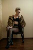 Attractive blonde model with pantyhose, black bra and fur coat posing provocatively on chair. Fashion portrait of sensual blonde, studio shot. Sensual female in black lingerie posing against wall. photo