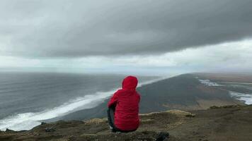 hombre sentar en rock pensativo Mira a atlántico Oceano ondas. famoso icónico acantilado punto de vista terminado Reynisfjara negro arena playa. persona mira para dirección y propósito en viajes video