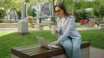 Carefree business woman sits outdoors with coffee and using laptop, remote work video