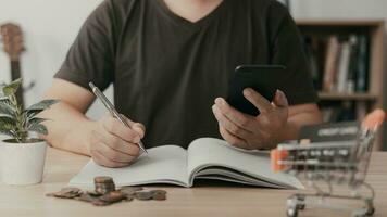 hombre utilizando teléfono inteligente y tomando notas en planificación. cuales es pago en línea, se sienta en el silla en el vivo habitación a hogar en un escritorio. el concepto de Finanzas y en línea compras. foto