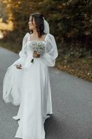 un hermosa morena novia en un blanco Boda vestir y velo con un Boda ramo. camina a lo largo el la carretera en el bosque. foto
