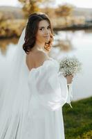 Portrait of a beautiful young bride in an elegant white dress with a long veil and a bouquet of gypsophila near the lake in soft light. The bride is looking at the camera. Portrait of the bride. photo