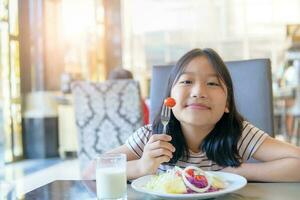 asiático linda pequeño niña comiendo Fresco tomate y ensalada en Mañana. foto