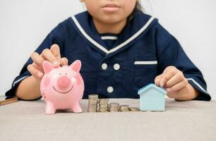 asiático niña poniendo monedas a cerdito banco y pequeño casa en blanco antecedentes foto