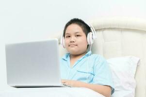 Obese schoolboy sitting and wear headphones to learn online from a laptop on their bed. photo