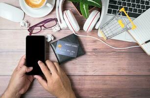 man touch on black screen mobile phone with credit card on wallet and laptop on wooden photo