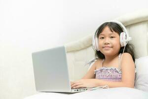 contento linda niña vistiendo auriculares a aprender en línea desde un ordenador portátil en su cama. foto