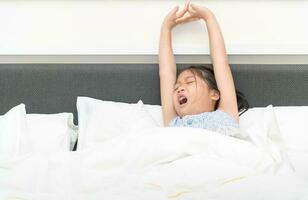 asiático linda niña despierta arriba y extensión en cama en mañana, foto