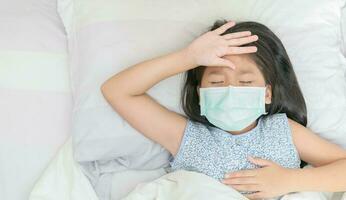 asiático niña vestir quirúrgico máscara tiene un alto fiebre y dolor de cabeza. foto