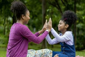 African American mother is laughing while playing patty cake with her young daughter while having a summer picnic in the public park for wellbeing and happiness concept photo