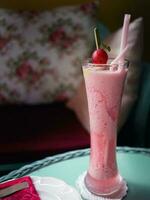 Cold red fruit juice in a tall glass decorated with 2 straws and a cherry on a blue table, selective focus with dark background, flowers cafe concept photo
