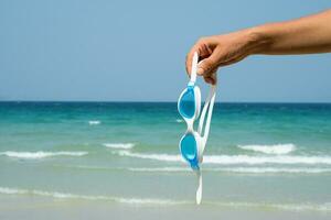 Hand holds swimming goggles agaist seascape. Tropical vacation concept. photo