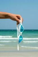 Hand holds swimming goggles agaist seascape. Tropical vacation concept. photo