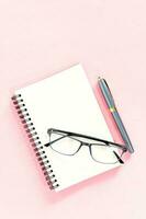 Flat lay of stationery with note book, pencil and eyeglasses on light pink background, Minimal work space. photo