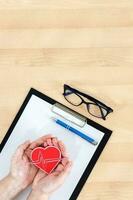 Female hands holding heart, clipboard and glasses on wooden desk. Top view. photo