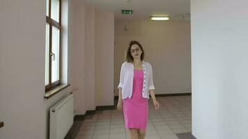 joven mujer en un brillante rosado vestir y blanco cárdigan caminando en un corredor video