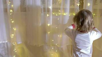 Little boy in christmas pajamas decorating curtain with christmas light beside the christmas tree video