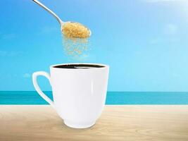 Spoon pouring granulated sugar into coffee mug on wooden table with the sea in the background photo