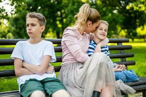 contento madre es sentado con su hijos en banco en parque. uno chico es ofendido y haciendo pucheros foto