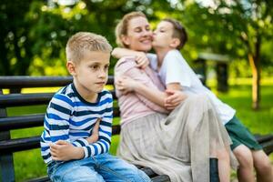 contento madre es sentado con su hijos en banco en parque. uno chico es ofendido y haciendo pucheros foto