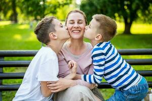 contento madre es sentado con su hijos en banco en parque. ellos son teniendo divertido juntos. Niños son besos su madre. foto