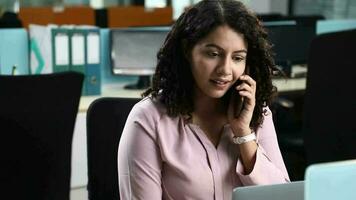 Video of smiling Young employee talking on cell phone while working on laptop