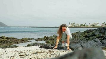 une fille dans blanc sur une volcanique plage touche cailloux. video