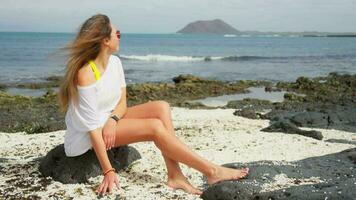 A girl in white sits on a volcanic shore, white small stones under her feet. video