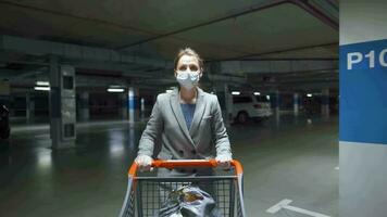 Woman in a medical mask and protective gloves walks with a grocery cart through an underground parking lot. Purchase during Covid-19 coronavirus pademia video