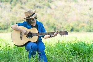 las mujeres de pelo corto usan sombrero y gafas de sol sentadas tocando la guitarra en el campo de hierba foto