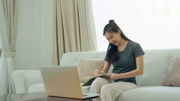 Asian woman sits on the sofa while taking notes from her computer screen while working or studying online at home. photo