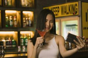 Asian woman taking a photo of herself while drinking whiskey at the bar.