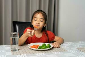 asiático pequeño niña comiendo sano vegetales con saborear. foto
