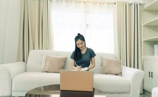 Asian woman sits on the sofa while taking notes from her computer screen while working or studying online at home. photo