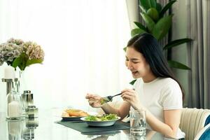 mujer joven asiática sonriendo mientras saca una ensalada en un plato y come felizmente en casa. foto