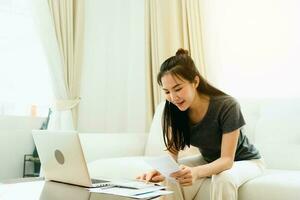 Young asian woman sits in the living room using a calculator to calculate family income and expenses and writes it down in his notebook or laptop at home. photo