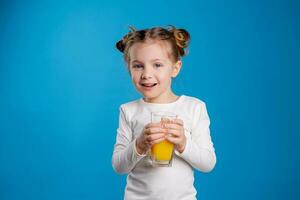 retrato de un pequeño niña de eslavo apariencia Bebiendo naranja jugo en un azul antecedentes foto