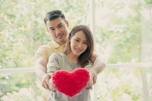 Happy couple sitting on the sofa and being a man embracing his girlfriend with love in the living room and smile and holding a red heart. Concept of romantic on valentine day. Proposal and marriage photo