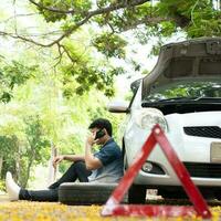 Asian man sitting beside car and using mobile phone calling for assistance after a car breakdown on street. Concept of vehicle engine problem or accident and emergency help from Professional mechanic photo