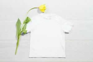 White children t-shirt mockup. Template blank kids shirt top view. White wooden background. Mother women day holiday. Yellow tulip. Baby tshirt with birthday flower. Spring look. photo